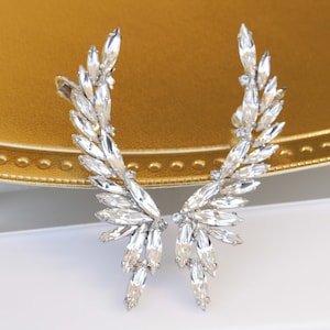 Pode incluir: Um par de brincos de cristal prateado em forma de asas. Os brincos são feitos de cristais transparentes e têm um design delicado e feminino.