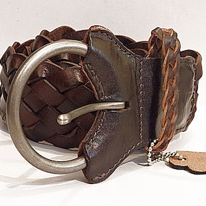 May include: A brown leather belt with a braided design and a silver buckle. The belt has a small leather tag attached to it.
