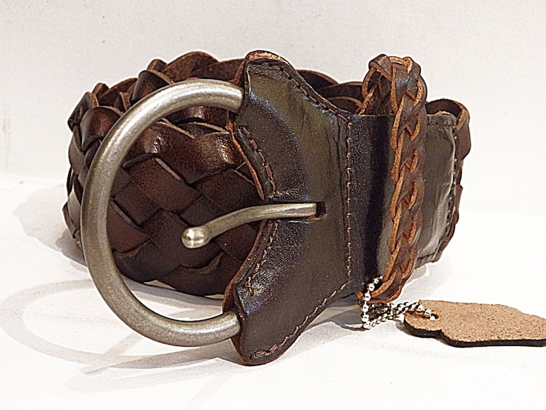 May include: A brown leather belt with a braided design and a silver buckle. The belt has a small leather tag attached to it.