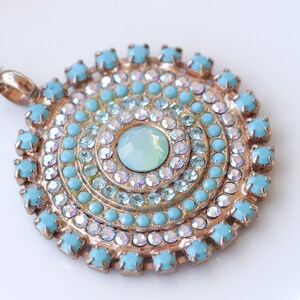 May include: A gold necklace with a large, round pendant featuring a turquoise and clear crystal design. The pendant is made up of concentric circles of turquoise and clear crystals, with a larger, clear crystal in the center.