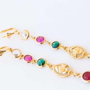 Könnte beinhalten: Ein Paar goldene Ohrringe mit einem keltischen Knotendesign. Jeder Ohrring hat einen weißen, rosa, grünen und roten Edelstein.