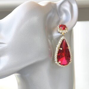 May include: A pair of gold tone teardrop earrings with a large red crystal and smaller red crystal accents. The earrings are set with clear rhinestones.