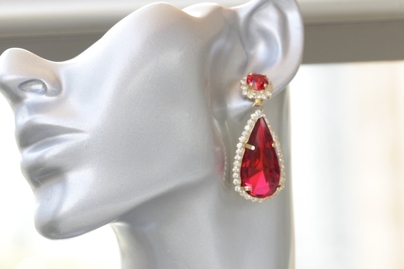 May include: A pair of gold tone teardrop earrings with a large red crystal and smaller red crystal accents. The earrings are set with clear rhinestones.
