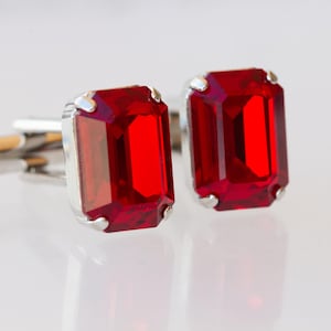 May include: Silver cufflinks featuring large, rectangular, ruby-red gemstones. The gemstones are set in a silver frame, and the cufflinks have a classic design. The background is a bright, reflective surface.