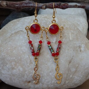May include: Gold dangle earrings with red and green glass beads and a treble clef charm. The earrings feature a large red round stone.