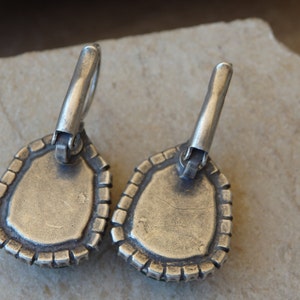 May include: A pair of silver earrings with a teardrop-shaped design. The earrings have a textured edge and a silver hook closure.