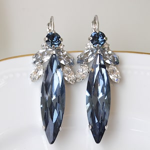 Puede incluir: Un par de pendientes colgantes con un cristal azul en forma de lágrima y cristales transparentes más pequeños. Los pendientes son plateados.