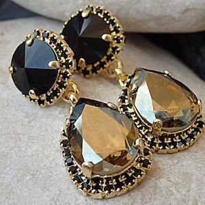 May include: A pair of gold tone dangle earrings with black and brown teardrop shaped crystals. The crystals are set in a gold tone metal frame with black beads around the edges.