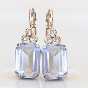 May include: A pair of silver-toned leverback earrings. Each earring features a light blue, rectangular gemstone with a faceted cut, accented by three smaller clear crystals above. The earrings are set against a white background.