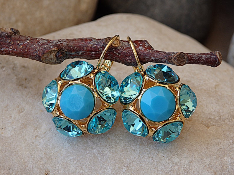 Puede incluir: Un par de pendientes de tono dorado con detalles de cristal azul. Los pendientes tienen forma de flor con un gran cristal azul en el centro.