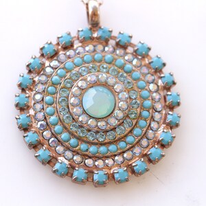 May include: A round pendant necklace with a turquoise and clear crystal design. The pendant features a central turquoise stone surrounded by concentric circles of smaller turquoise and clear crystals.