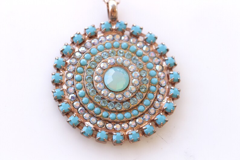 May include: A round pendant necklace with a turquoise and clear crystal design. The pendant features a central turquoise stone surrounded by concentric circles of smaller turquoise and clear crystals.