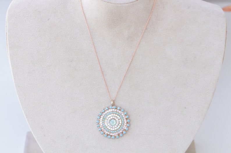 May include: A rose gold chain necklace with a round pendant featuring a concentric circle design of light blue and white rhinestones.
