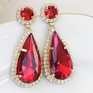 May include: A pair of dangle earrings with large teardrop-shaped red crystals surrounded by a halo of smaller clear crystals. The earrings have gold-toned metal settings.