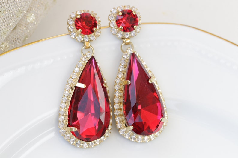 May include: A pair of dangle earrings with large teardrop-shaped red crystals surrounded by a halo of smaller clear crystals. The earrings have gold-toned metal settings.