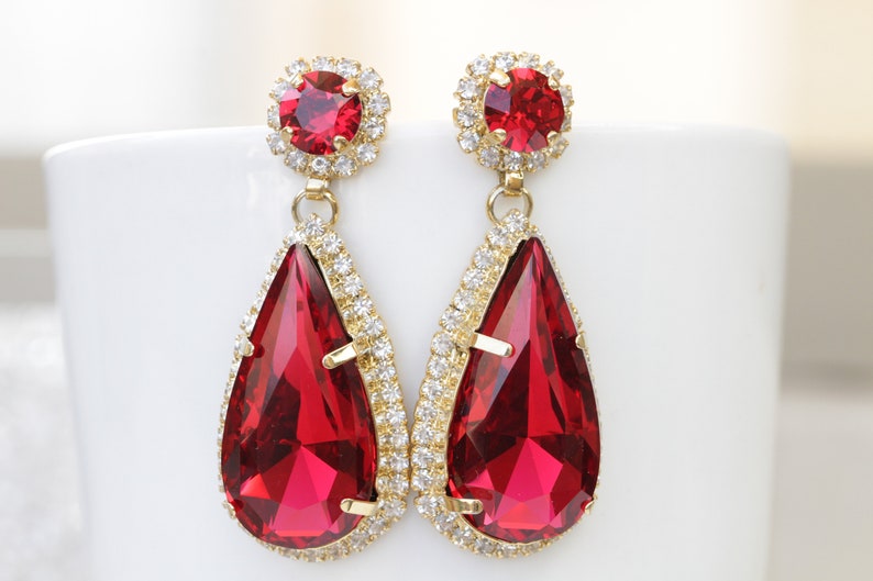 May include: A pair of gold tone dangle earrings with teardrop shaped red crystal stones and smaller round red crystal stones at the top.