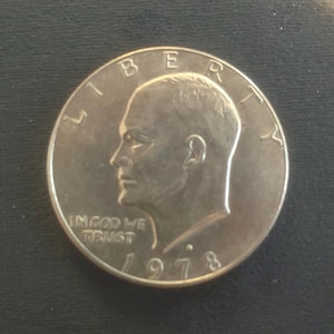 Puede incluir: Una moneda de un dólar Eisenhower de plata de 1973. La moneda presenta un perfil de un hombre, las palabras "LIBERTY" e "IN GOD WE TRUST", y el año "1973". La moneda es redonda y tiene un aspecto metálico brillante.