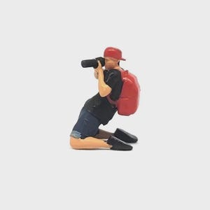 May include: Miniature figurine of a photographer kneeling, capturing an image. The figure wears a red cap, black shirt, denim shorts, and black shoes. A red backpack is visible. The photographer is holding a black camera.