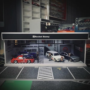 May include: A detailed diorama depicting a garage with model cars. The scene includes a red sports car, a white estate car, and a grey saloon. The sign above the garage reads "Rocket Bunny".