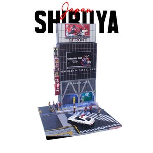 May include: A miniature diorama of the Shibuya district in Japan, featuring a multi-story building with advertisements for QFRONT, Chainsaw Man, and Starbucks Coffee. A police car is on the crosswalk, and the word "Shibuya" is displayed.
