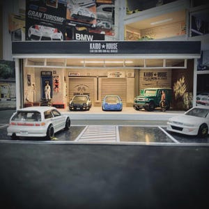 以下が含まれることがあります： 「KAIDO HOUSE」の文字が上部にある、ミニチュアの車のガレージのジオラマ。白いハッチバック、白黒の車、青い車、緑のSUVなど、いくつかのミニカーがガレージの前に駐車されています。緑のSUVの近くにフィギュアが立っています。