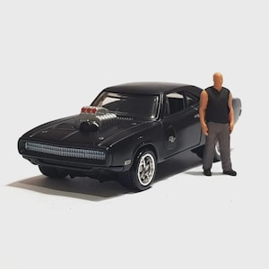 May include: A black die-cast model car with a large engine component and chrome wheels. A small figurine of a man in a black shirt and gray pants stands next to the car. The car has a classic muscle car design.