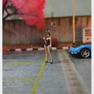 May include: Miniature figure of a woman in a black bunny costume, holding a bottle and a small container, standing on a gray asphalt surface. A blue sports car is parked nearby. The background includes a pink tree and a building.