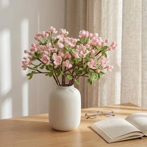 May include: A bouquet of crocheted light pink cherry blossom flowers with green leaves and brown stems arranged in a smooth, matte white ceramic vase. The floral arrangement features numerous small buds and open blossoms with intricate yarn textures.
