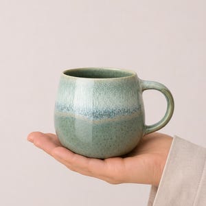 Könnte beinhalten: Ein abgerundeter Keramikbecher mit einer reaktiven Glasur in einem Farbverlauf aus Salbeigrün und sanften Blautönen. Die Oberfläche zeigt eine gesprenkelte Textur mit glattem, glänzendem Finish, einem geschwungenen Henkel und einer weiten Öffnung.