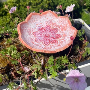Könnte beinhalten: Eine blütenförmige Keramikschale mit einem rosa-weißen Blumendesign. Die Schale ist von grünen Pflanzen und rosa Blüten umgeben. Die Schale wird wahrscheinlich für dekorative Zwecke oder als kleine Servierschale verwendet.