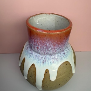 Könnte beinhalten: Eine handgefertigte Keramikvase mit einem einzigartigen Design. Der obere Teil ist rot-orange, geht in einen weißen und violetten Farbton über. Der Boden ist ein gesprenkeltes Beige mit weißer Glasur, die heruntertropft.