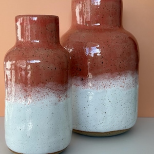 Könnte beinhalten: Zwei Keramikvasen mit zweifarbigem Design. Die obere Hälfte jeder Vase ist ein gesprenkeltes Rotbraun, das in eine gesprenkelte weiße Basis übergeht. Die Vasen haben eine abgerundete Form mit zylindrischem Hals.