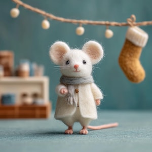 Puede incluir: Una figura de ratón blanco hecha a mano con una bufanda gris y un abrigo color crema. El ratón tiene patas rosas y una larga cola rosa. Un pequeño calcetín navideño decorativo cuelga en el fondo.