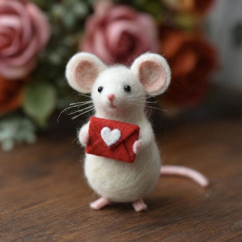 May include: A white felt mouse figurine holding a red envelope with a white heart. The mouse has large ears, a pink nose, and whiskers. The background is blurred with pink and orange flowers.