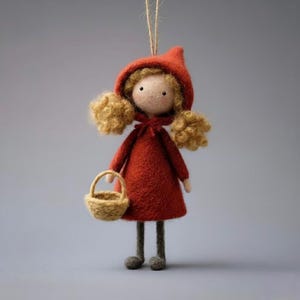 May include: A handmade felt doll dressed in a red hooded cloak and dress, holding a small woven basket. The doll has curly blonde hair and gray legs. A string is attached for hanging. The doll is approximately 6 inches tall.