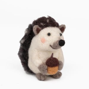 May include: A handcrafted felt hedgehog figurine with a white body, dark brown quills, and a black nose. The hedgehog holds a brown and tan acorn. The cheeks are a soft pink. A charming woodland creature.
