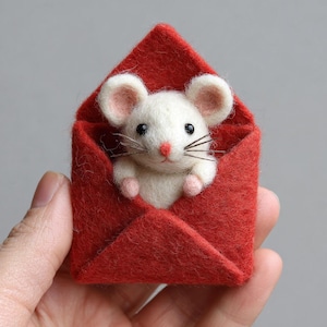 May include: A handmade felt mouse peeking out of a red felt envelope. The mouse is white with pink ears, paws, and nose, and has black eyes and whiskers. The envelope is a vibrant red.