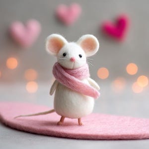 May include: A handcrafted white felt mouse wearing a pink scarf, standing on a pink felt surface. The mouse has large, round ears, a pink nose, and small black eyes. Soft pink felt hearts are in the background, with bokeh lights.
