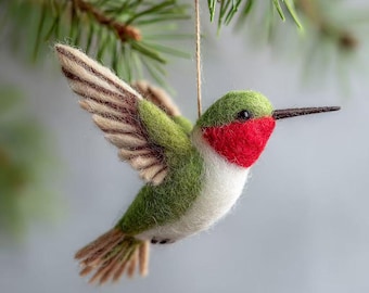 May include: A felt hummingbird ornament with outstretched wings, suspended from a natural twine string. The bird features a green back, red throat, white chest, and tan wings. The hummingbird is hanging from a green pine branch.