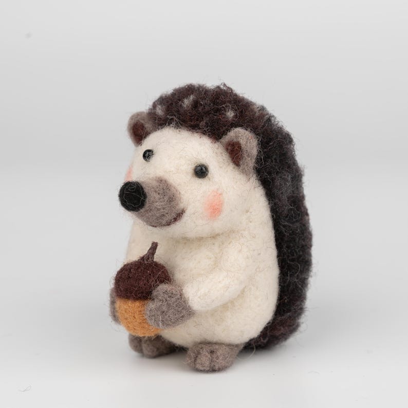 May include: A handmade felt hedgehog figurine with a white body, dark brown quills, and a black nose. The hedgehog holds a brown and orange acorn. The cheeks are a soft pink. This whimsical woodland creature is a charming decorative item.