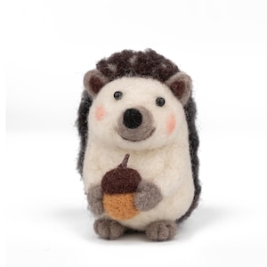 May include: A handcrafted felt hedgehog figurine with a white body, dark brown quills, and a smiling face with pink cheeks. The hedgehog is holding a brown and tan acorn, perfect for fall decor.
