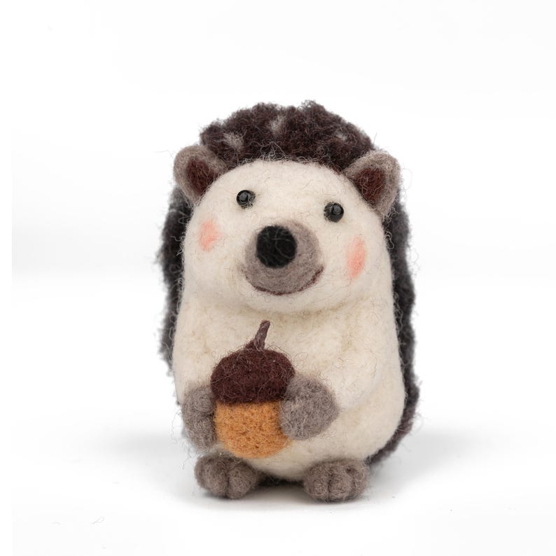 May include: A handcrafted felt hedgehog figurine with a white body, dark brown quills, and a smiling face with pink cheeks. The hedgehog is holding a brown and tan acorn, perfect for fall decor.