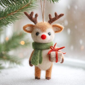 Puede incluir: Adorno navide&ntilde;o artesanal de un reno con cuernos marrones, nariz roja y bufanda verde. El reno sostiene un peque&ntilde;o regalo envuelto en papel blanco y atado con una cinta roja. El adorno est&aacute; suspendido por una cuerda.