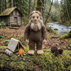 Può includere: Una figura artigianale in feltro di Bigfoot è in piedi su un tronco muschioso con un mini campeggio. La figura marrone ha un viso chiaro e capelli ispidi. Una piccola tenda, un sacco a pelo arrotolato e un falò sono nelle vicinanze, con una piccola capanna sullo sfondo.