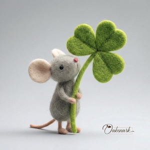 May include: A handmade felt mouse figure in grey, holding a large green four-leaf clover. The mouse has pink ears, nose, and feet. The clover's stem is green, set against a soft grey background.