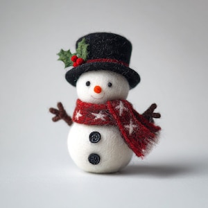 Peut inclure: Figurine de bonhomme de neige artisanal avec un chapeau haut de forme noir et une écharpe rouge à motifs d'étoiles blanches. Il a des bras en brindilles, un nez en carotte, des yeux en boutons et une bouche souriante. Le chapeau est orné d'une branche de houx.
