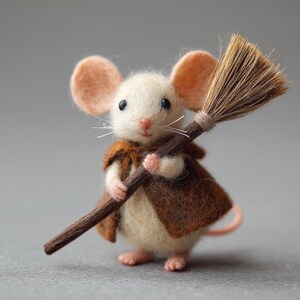 May include: A handcrafted felt mouse figurine, wearing a brown cape and holding a miniature broom. The mouse has cream-colored fur, large pink ears, and tiny pink paws. The broom is made of brown bristles and a wooden handle.