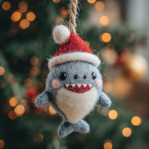 Puede incluir: Adorno navideño hecho a mano con forma de tiburón, con un gorro de Papá Noel rojo y blanco. El tiburón es gris con vientre blanco, mejillas rosas y boca roja. Colgado de una cuerda de hilo natural, ideal para la decoración navideña.