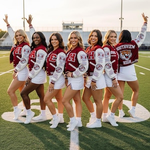 Może przedstawiać: Grupa cheerleaderek w bordowych i białych kurtkach typu letterman i białych spódnicach. Kurtki mają naszywki na rękawach i napis "Southview High" na plecach. Stoją na zielonym boisku do piłki nożnej.