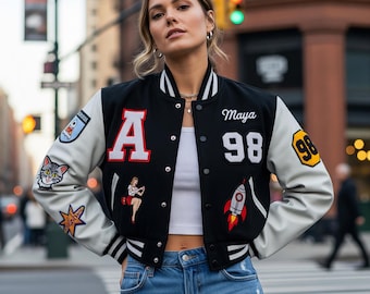 May include: Black varsity jacket with white sleeves, featuring the letter "A" and the number "98" in white. The jacket has various patches, including a cat, a rocket, and a pin-up girl. The name "Maya" is embroidered on the jacket.